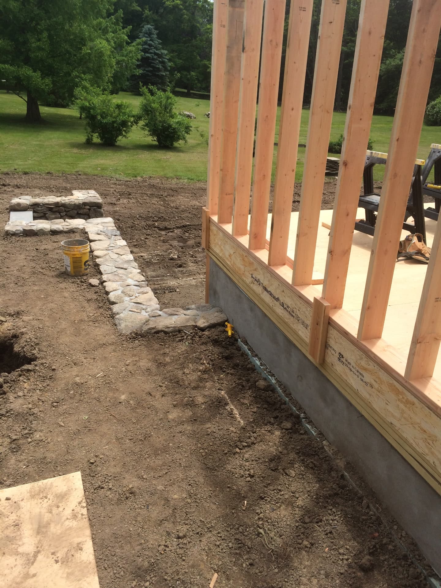 New wall framing rising on a foundation beside a hand-laid fieldstone wall